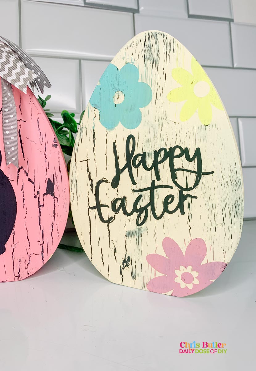 a cream colored crackled wooden egg with a blue flower, yellow flower, pink flower and the words "Happy Easter" in charcoal 